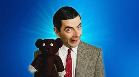 Man in beige blazer and red tie holding a brown teddy bear puppet against a blue background